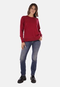 Pullover rosso a maniche lunghe con collo a giro, texture morbida e orlo a coste, abbinato a jeans blu aderenti e stivali neri con lacci.