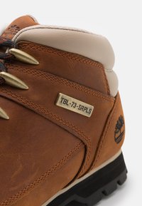 Botte de randonnée en cuir marron avec des éléments en laiton, semelle noire et col rembourré beige. Présente une étiquette en métal gravée "TBL-73-SRPLS".