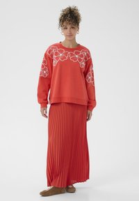 Femme aux cheveux bouclés portant un sweat-shirt floral rouge, une longue jupe plissée rouge, des baskets marron et des boucles d'oreilles en or, se tenant sur fond blanc.
