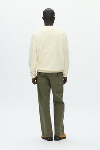 Homme debout, vu de dos, portant un sweat-shirt crème, un pantalon vert olive avec des poches arrière boutonnées, et des chaussures marron avec des semelles noires.