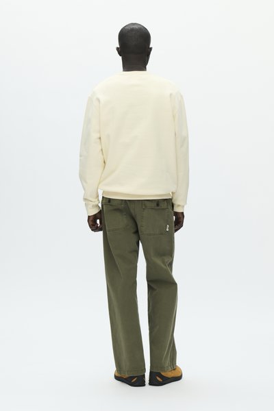 Homme debout, vu de dos, portant un sweat-shirt crème, un pantalon vert olive avec des poches arrière boutonnées, et des chaussures marron avec des semelles noires.