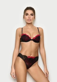 Kvinde iført sort og rød blonde lingeri sæt, med underwire bh og matchende højt skårne trusser, poserende mod en ensfarvet lys baggrund.