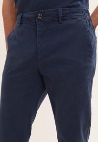 Pantalone od pamuka u mornarsko plavoj boji s ravnim prednjim dijelom, zatvaranjem na dugme, petljama za remen i bočnim džepovima. Glatka tekstura i kroj prilagođen tijelu.