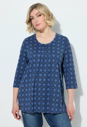 Femme aux cheveux blonds courts portant une tunique bleue à motifs à manches longues et un jean bleu, debout sur un fond blanc.