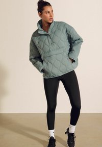 Lys blå quiltet pullover med høj krave, trykknaplukning og frontlomme. Sammen med sorte leggings og sportssko.