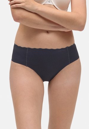 Mujer vestida con sujetador beige y braguitas azules marino sin costuras con bordes en forma de ola, de pie con los brazos cruzados sobre el pecho.
