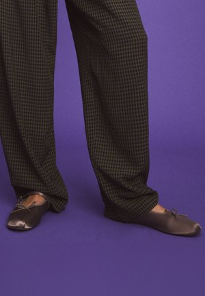 Personne portant un pantalon ample à carreaux et des ballerines sombres, debout sur un sol violet avec un fond violet.