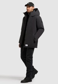 Giacca nera isolante con colletto alto e grande cappuccio, dotata di zip frontale e tasche laterali. Indossata con jeans neri e stivali.