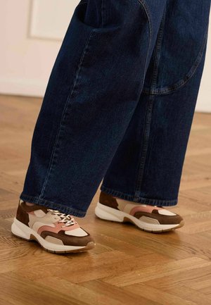 Voeten met sneakers in meerdere tinten bruin, beige en roze, gecombineerd met wijde donkerblauwe jeans, staand op een houten parketvloer.