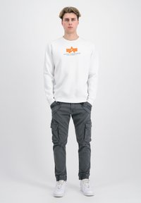 Hvid sweatshirt med orange logo, omgivet af grå cargo bukser med sidelommer, og hvide sneakers. Enkel baggrund.