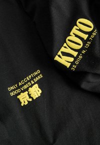 Sort bomuldsstof med gul tekst. Indeholder ordene "KYOTO" og "ONLY ACCEPTING GOOD VIBES & SAKE" med japanske tegn i fed typografi.