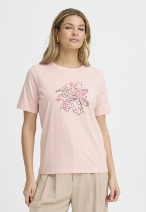 Kvinde iført en lyserød T-shirt med et blomstermønster i rødt og brunt, kombineret med beige plisserede bukser, stående mod en hvid baggrund.