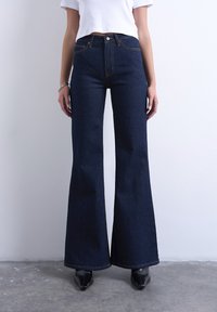 Άτομο που φοράει ψηλόμεσα σκούρα μπλε καμπάνα τζιν, λευκό κοντό μπλουζάκι, μαύρα μυτερά παπούτσια, στέκεται σε τσιμεντένιο δάπεδο μπροστά σε λευκό τοίχο.