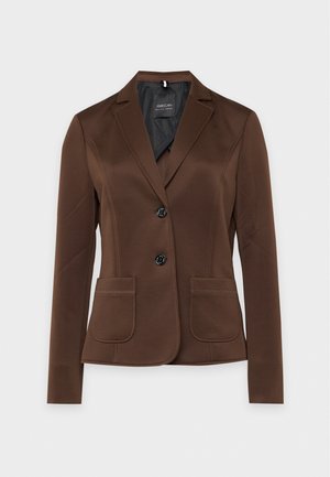 Une femme porte un blazer marron, un chemisier crème, un pantalon à motifs et des chaussures à talons marron devant un fond de studio uni.