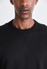 Svart crewneck hettegenser laget av bomull med teksturert søm detalj på kragen og skulderen. Enkel, udekorerte design.