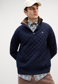 Quilted sötétkék pulóver magas gallérral és barna kordbársony bélés, elülső patentos zárással és logóval. Kockás inggel rétegezve.