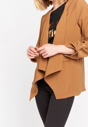 Femme portant une veste drapée couleur camel sur un t-shirt graphique noir et un pantalon noir, mains doucement jointes devant.