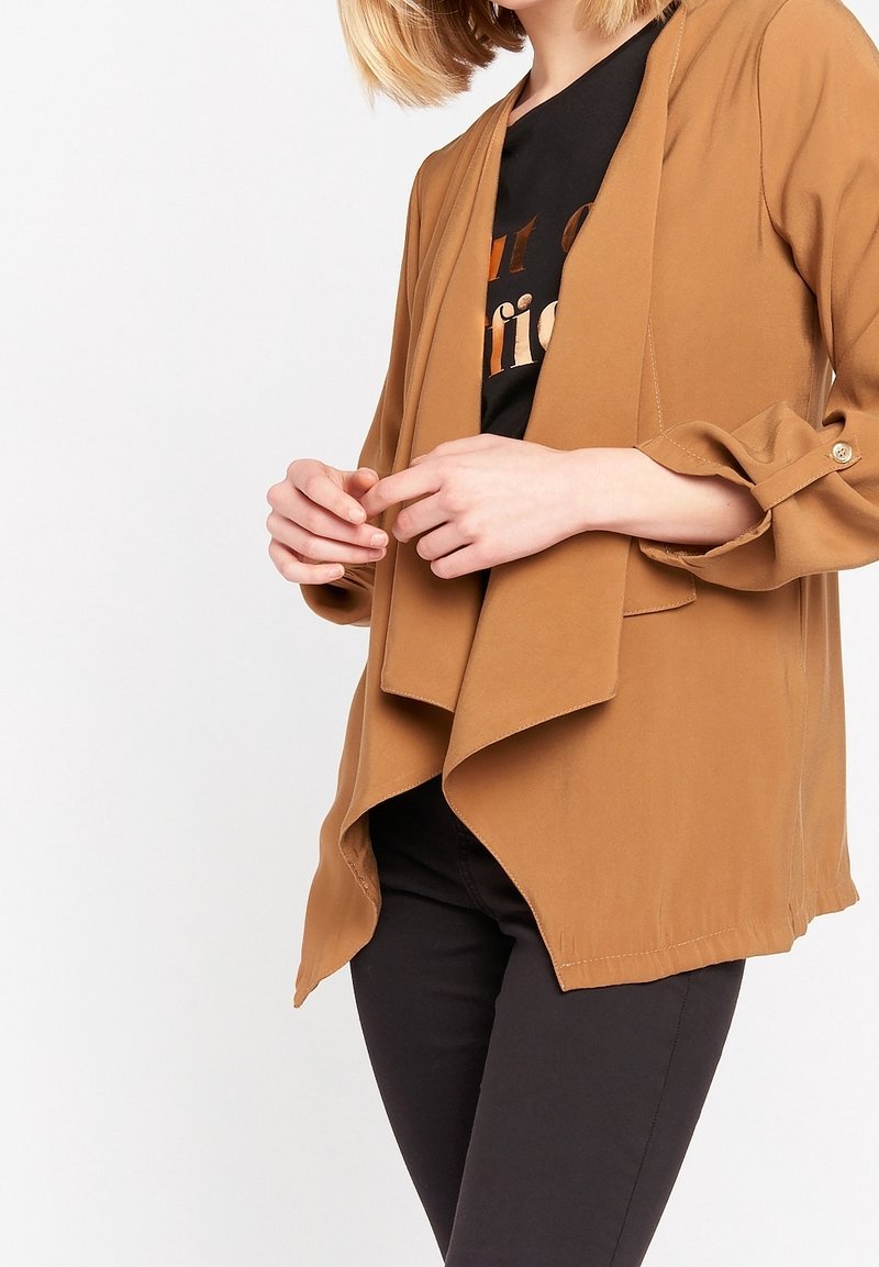 Femme portant une veste drapée couleur camel sur un t-shirt graphique noir et un pantalon noir, mains doucement jointes devant.