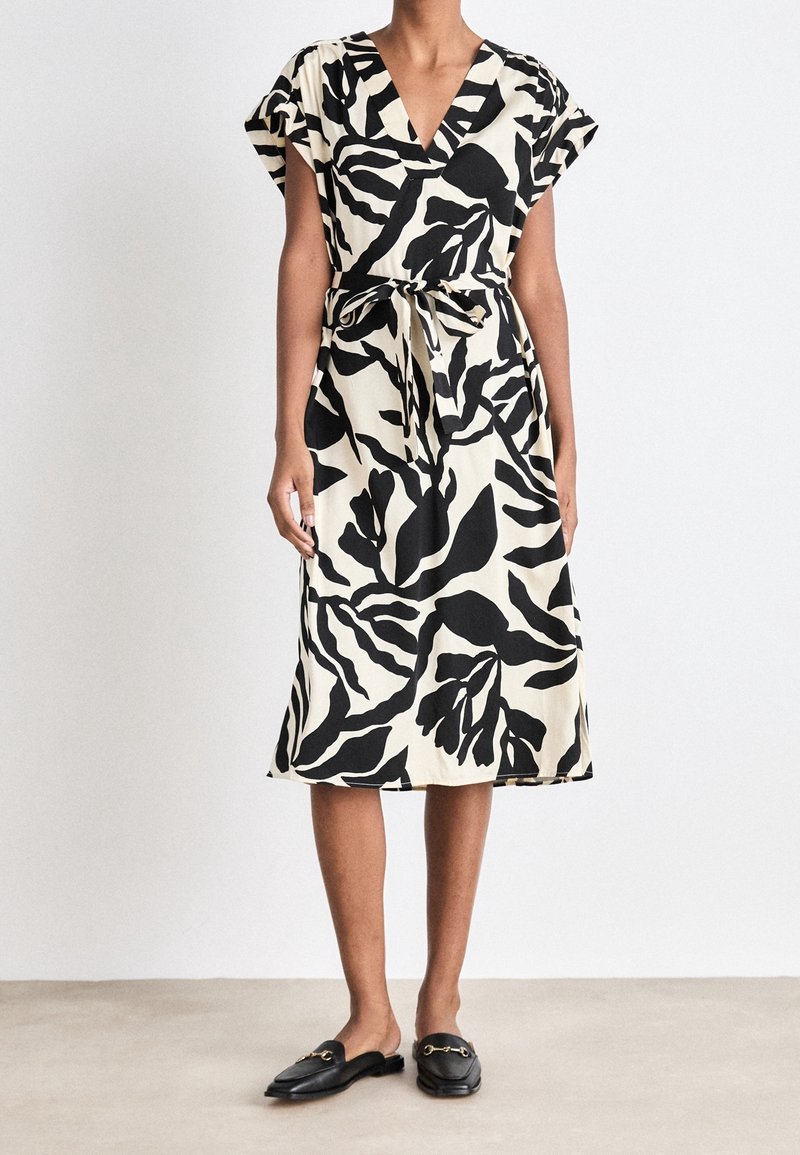 Femme portant une robe portefeuille florale noire et crème à manches courtes, avec une taille cintrée, et des mocassins noirs à enfiler, debout sur un sol neutre.