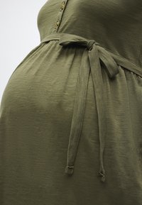 Κοντινή όψη ενός ελιάς-πράσινου φορέματος εγκυμοσύνης με δεμένη ζώνη στη μέση και τρία κουμπιά στο στήθος.