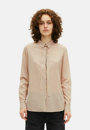 Vrouw met krullend haar, gekleed in een beige blouse met rode stippen en zwarte broek, staand tegen een effen witte achtergrond.