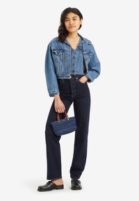 Mlada ženska, oblečena v skrajšano jeans jakno in temne hlače z visokim pasom, drži majhno jeans torbico z rjavimi ročaji.