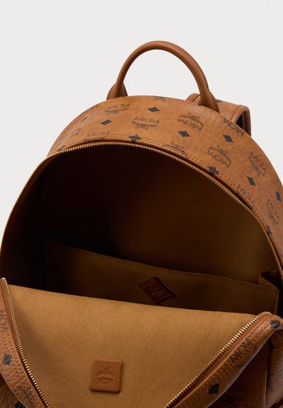 Brown leather backpack with black MCM logos, open to show empty interior with a flat pocket and gold zipper detail.
