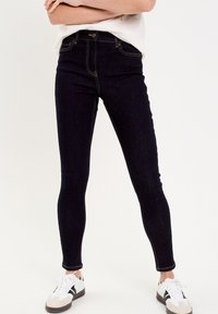 Jean skinny en denim noir avec taille haute, poches avant, couture jaune et fermeture par bouton. Associé à des baskets noires et blanches.