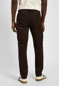 Brązowe bawełniane chinosy o prostym kroju, z tylnymi kieszeniami zapinanymi na guziki oraz czystym wykończeniem, w połączeniu z białymi i granatowymi butami w paski.