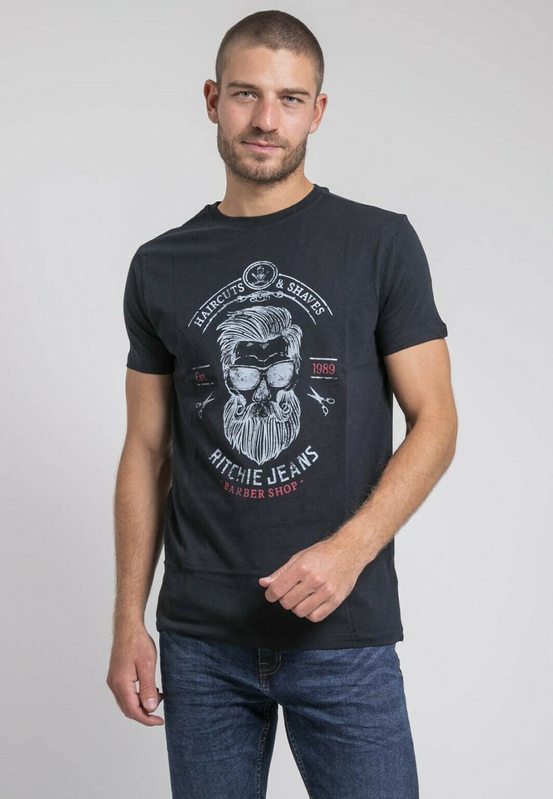 T-shirt en coton noir avec un imprimé graphique d'un homme barbu portant des lunettes de soleil, accompagné de texte rouge et blanc évoquant le thème d'un salon de coiffure.
