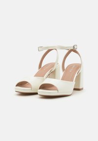 Weiße patentierte Leder-High Heels mit offener Zehe, breitem Riemen über dem Fuß, Knöchelriemen und Blockabsatz. Glatte Textur und minimales Design.
