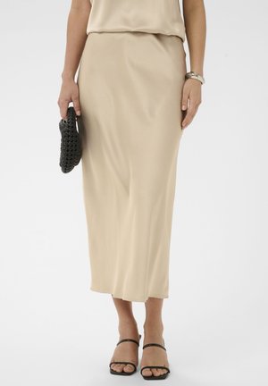 Femme portant une jupe midi beige et un haut sans manches, tenant une pochette noire tressée, et portant des sandales noires à lanières ainsi qu'un bracelet argenté.