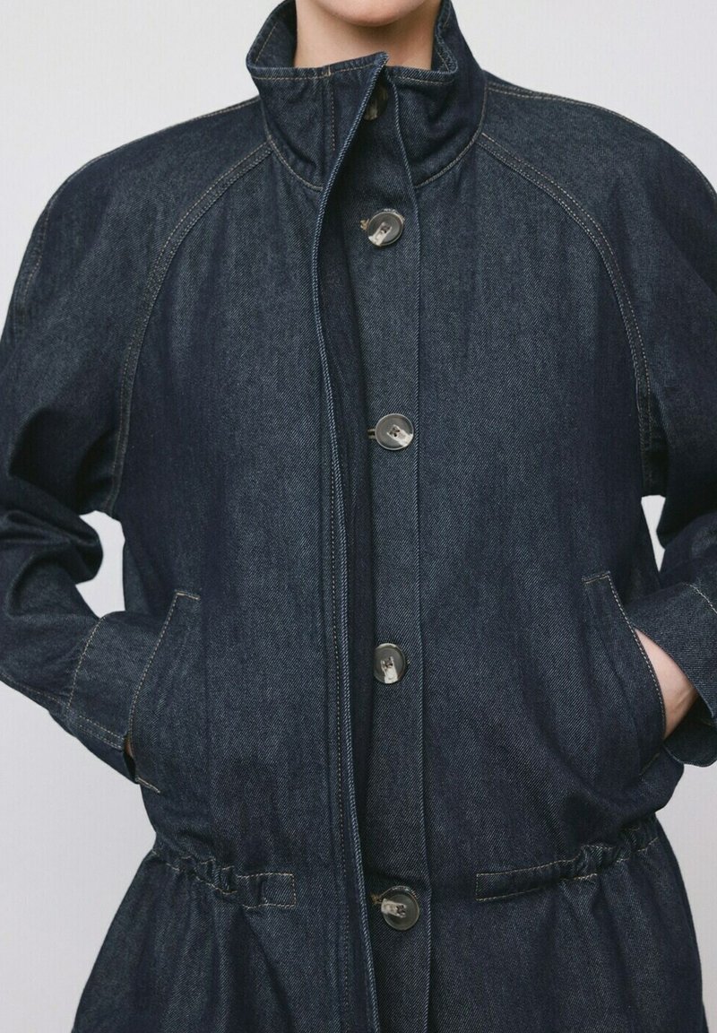 Personne portant une veste en denim bleu foncé avec de gros boutons, un col haut, des poches latérales et une taille froncée, les mains partiellement dans les poches.