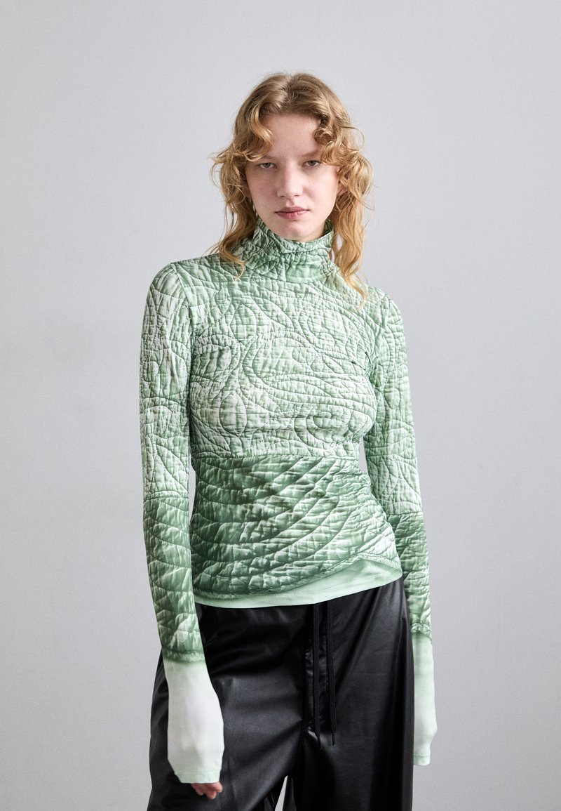 Long-sleeve, green, quilted top with a high collar. Features a textured pattern and fitted design. Paired with loose black pants.