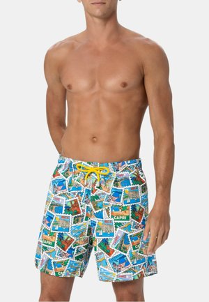 Uomo senza maglietta che indossa pantaloncini da bagno colorati con disegni di cartoline di viaggio e un laccetto giallo, in piedi con una mano al fianco.