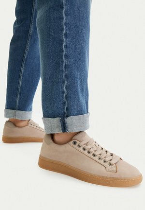 Nohy v béžových teniskách na tkaničky s gumovými podrážkami a vyhrnutými modrými džíny na bílém pozadí.