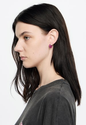 Orecchini a cerchio viola con una finitura liscia e piccole pietre decorative, indossati da un modello con lunghi capelli scuri, davanti a uno sfondo bianco.