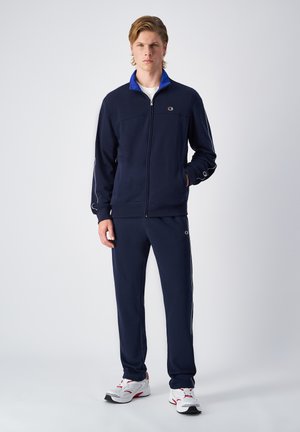Navy tracksuit featuring a zip-up jacket with a blue collar and pants with white side stripes. Material appears soft, designed for comfort.