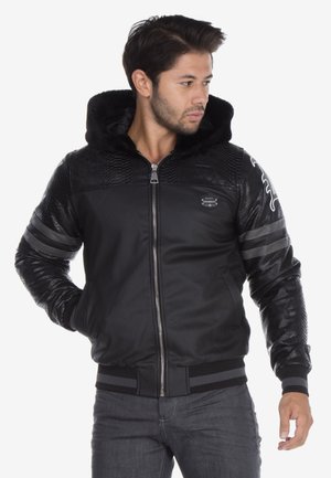 Veste bomber noire avec capuche en fausse fourrure, accents de panneaux texturés, deux rayures horizontales sur les manches et fermeture éclair. Présente un design ajusté.