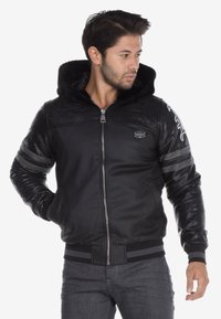 Schwarze Bomberjacke mit einer Kapuze aus strukturiertem Strick, Reißverschluss und kontrastierenden, glatten Leder-Schulterpaneelen. Mit grauen Streifen an den Ärmeln ausgestattet.