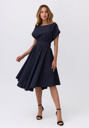 Kobieta w ciemnoniebieskiej sukience o wzorze do kolan z szerokim paskiem w talii oraz czarnych szpilkach na kostkę, stojąca na jednolitym tle.