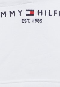 Tessuto in cotone bianco con il testo "TOMMY HILFIGER" e "EST. 1985" ricamato in blu marin, con un piccolo dettaglio del logo rosso, bianco e blu.