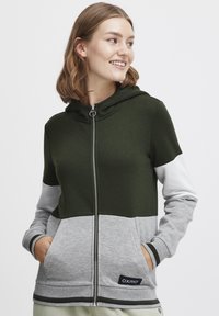 Huvtröja med dragkedja som har paneler i grönt, grått och vitt, utrustad med sidofickor och en märkt patch på den nedre fållen.