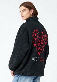 Jeune personne portant une veste polaire noire avec un motif de cœurs rouges en grappes et le texte "Sweet Heart" au dos, associée à un jean bleu clair.