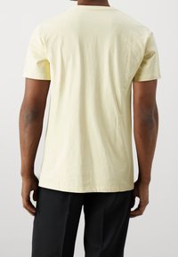 Žluté bavlněné tričko s krátkými rukávy a kulatým výstřihem, pohled zezadu. Hladká textura, minimalistický design, žádné viditelné vzory ani akcenty.