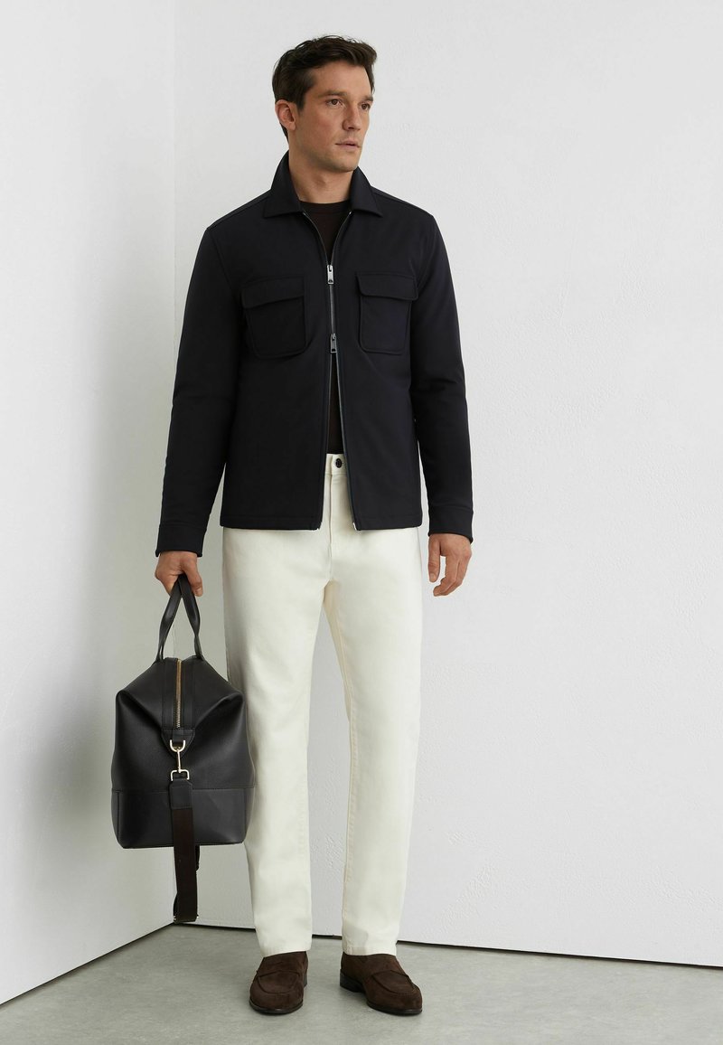 Férfi fekete cipzáras dzsekiben, fehér nadrágban, barna cipőben, fekete bőr sporttáskát tartva, fehér fal előtt áll.