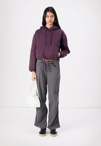 Cropped hoodie viola con cordino, pantaloni ampi grigi e borsa bianca. Scarpe nere con suole chiare. Sfondo neutro.