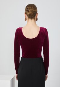 Bordo aksominis bodysuit'as su ilgomis rankovėmis ir plačiu apykaklės iškirpimu, derinamas su juoda padidinta juosmens suknele, esantis šviesiame fone.
