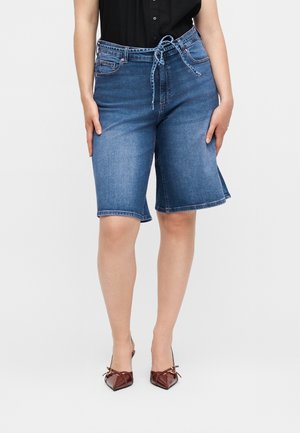 Kvinde iført blå knælange denimshorts med bundet bælte, sort ærmeløs skjorte og brune spidse hæle.