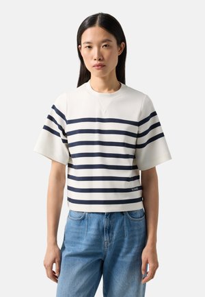 Kvinde med langt mørkt hår iført en hvid kortærmet skjorte med marineblå vandrette striber og lyseblå jeans, stående mod en ensfarvet baggrund.